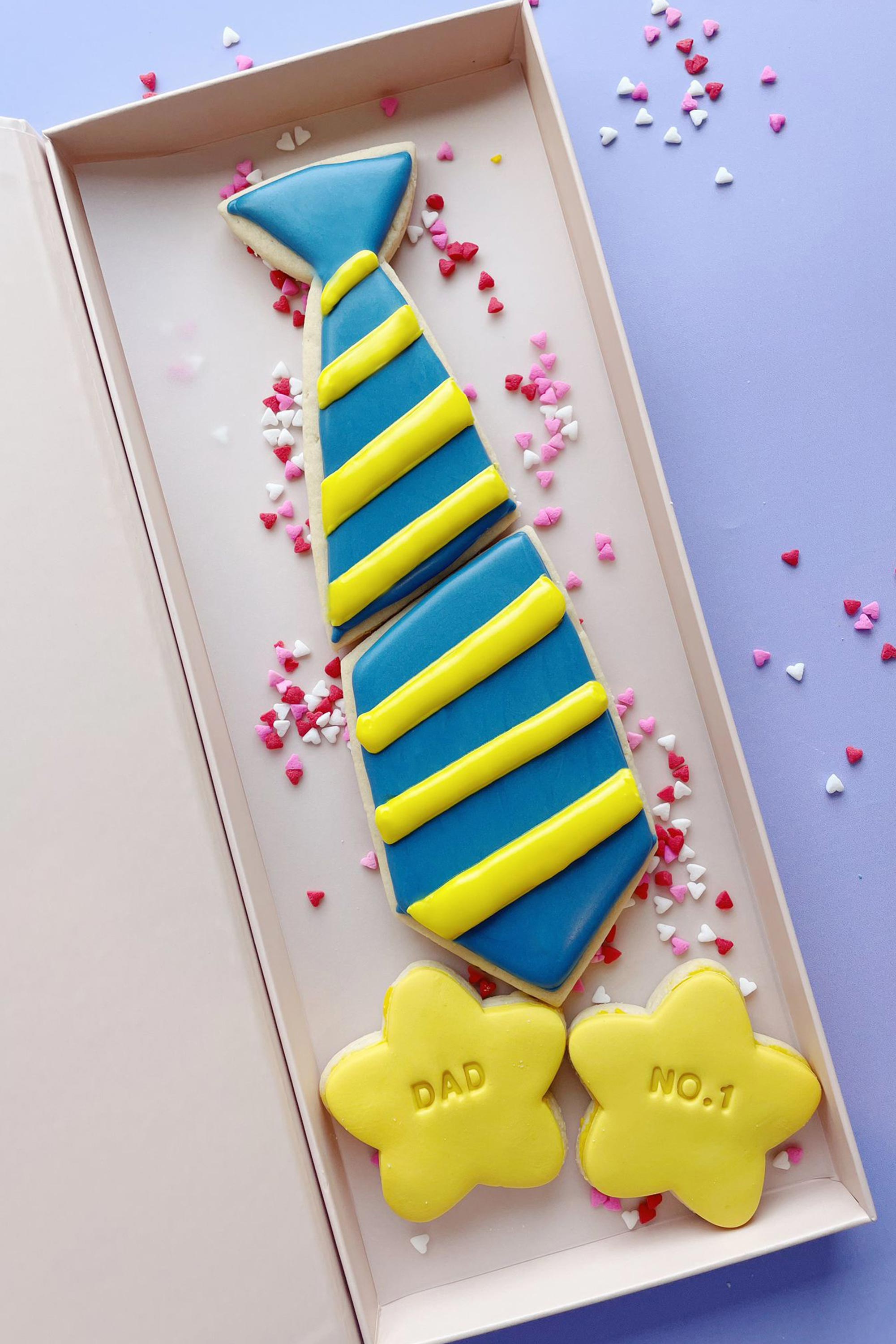 Decorative cookies shaped like a tie and stars with 'DAD' and 'NO.1' text on a box against a purple background.