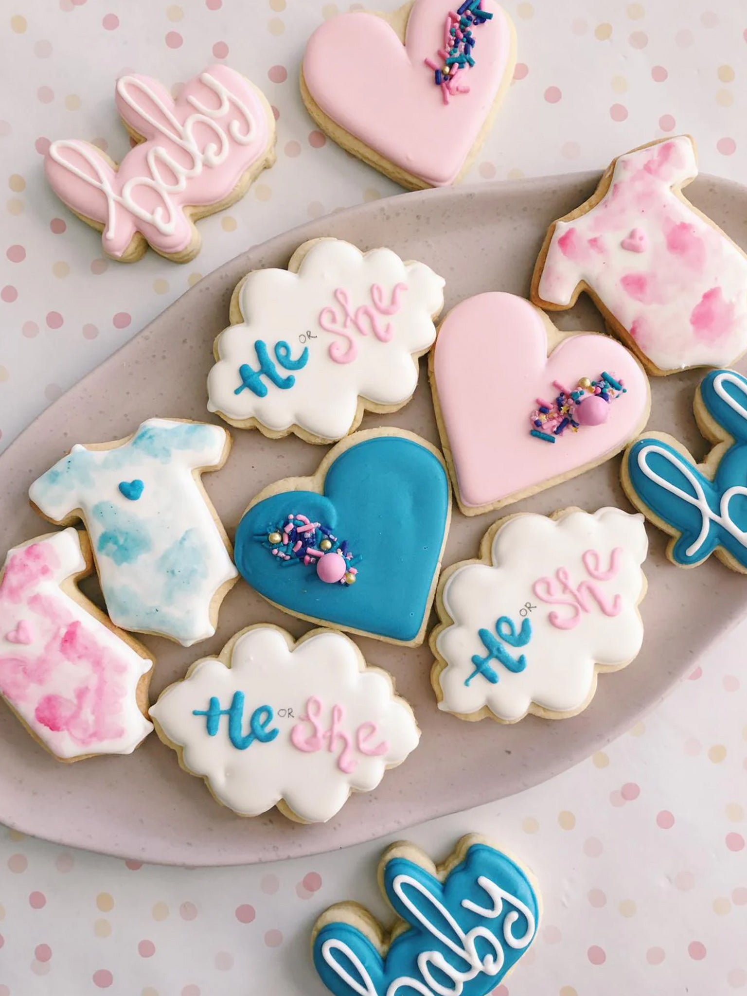 A variety of decorated sugar cookies with pink and blue themes, featuring the words 'baby' and 'he or she' on them, along with heart and cloud shapes.